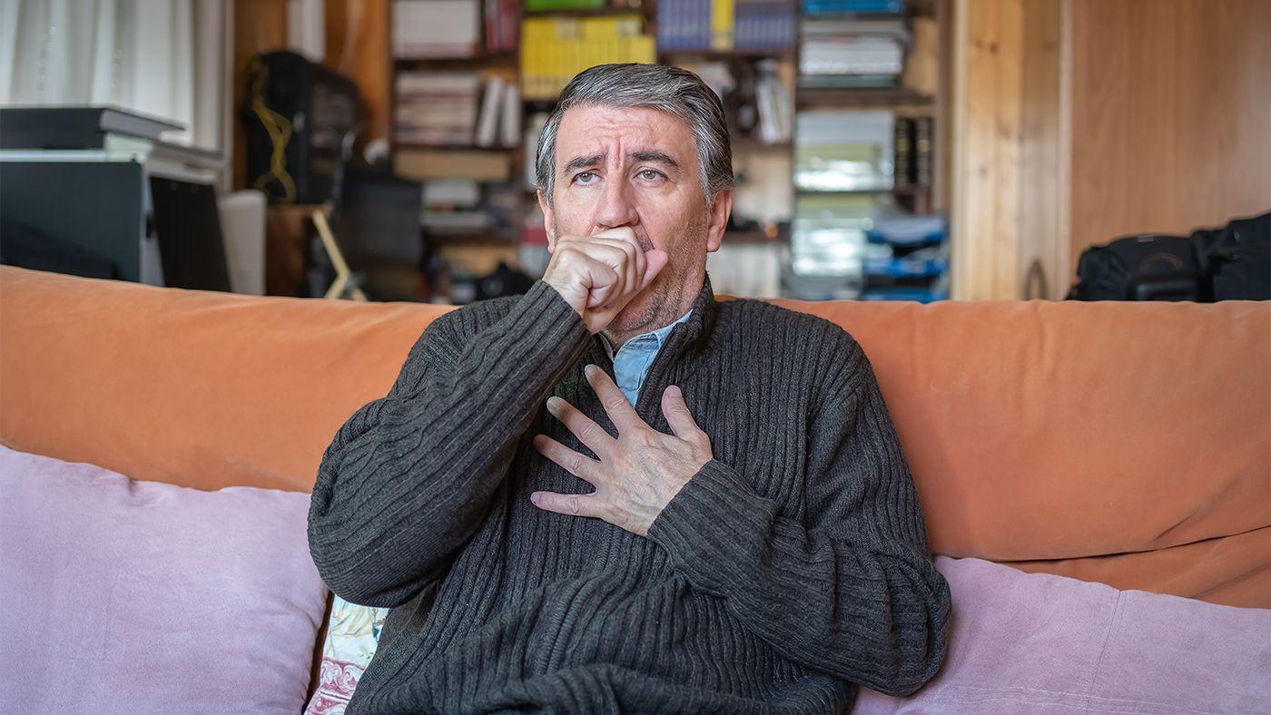 Elderly man coughing into his hand while sitting on a couch, representing the need for over-the-counter cough relief. 